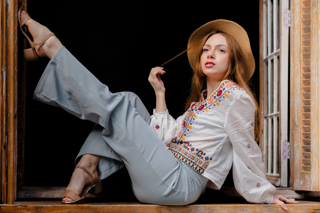 Young woman posing elegantly by a windowsill, wearing fashionable embroidered blouse and hat.
